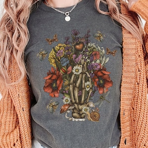 May include: Dark gray t-shirt featuring a detailed graphic of a skeleton hand holding a bouquet of flowers. The bouquet includes red poppies, daisies, and other colorful blooms, with butterflies and a snail. The shirt is a comfortable, casual style.