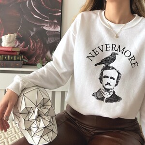 May include: White sweatshirt with a black and white illustration of Edgar Allan Poe with a raven perched on his head. The text "NEVERMORE" is printed in a black arc around the illustration.