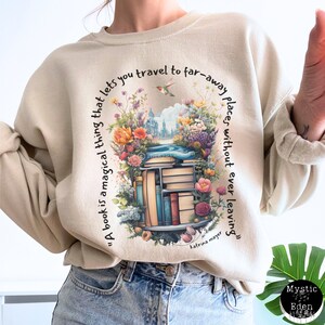 K&ouml;nnte beinhalten: Ein beiger Pullover mit einem Zitat &uuml;ber B&uuml;cher, das auf der Vorderseite aufgedruckt ist. Das Zitat lautet: "Ein Buch ist eine magische Sache, die es dir erm&ouml;glicht, an ferne Orte zu reisen, ohne jemals zu gehen." Das Zitat wird Katrina Mayer zugeschrieben. Der Pullover zeigt eine farbenfrohe Illustration eines B&uuml;cherregals mit B&uuml;chern und Blumen.