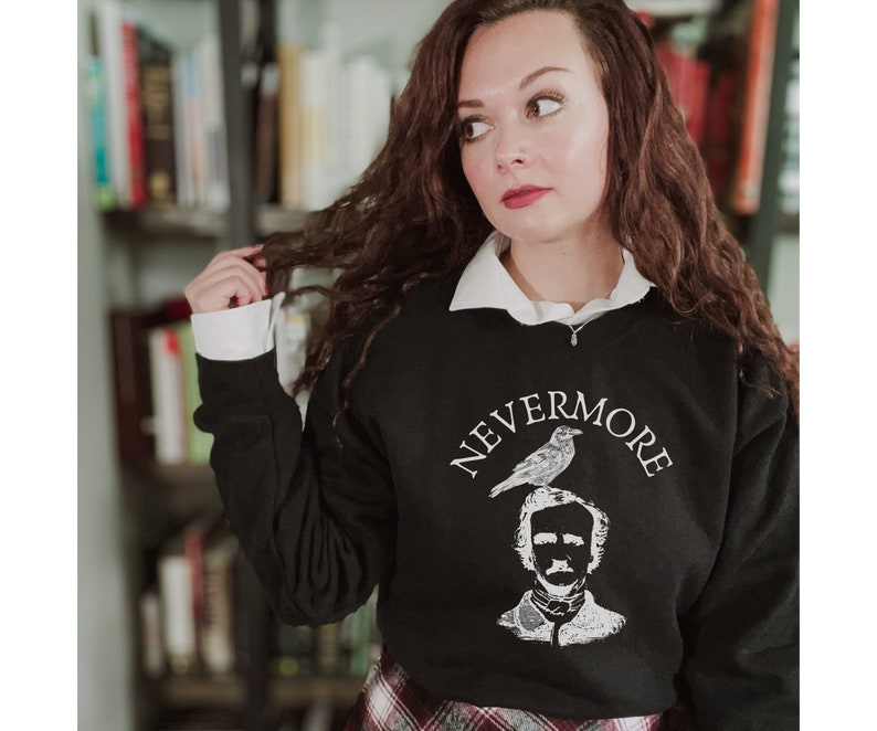 May include: A black sweatshirt with a white graphic print of Edgar Allan Poe with a raven perched on his head. The text "NEVERMORE" is printed in a white arc above the image. The model is wearing a white collared shirt underneath the sweatshirt.