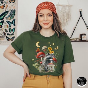 May include: A green t-shirt with a graphic of a raccoon standing in a forest scene with mushrooms, butterflies, and stars. The raccoon is looking to the right. The shirt has a crew neck and short sleeves.