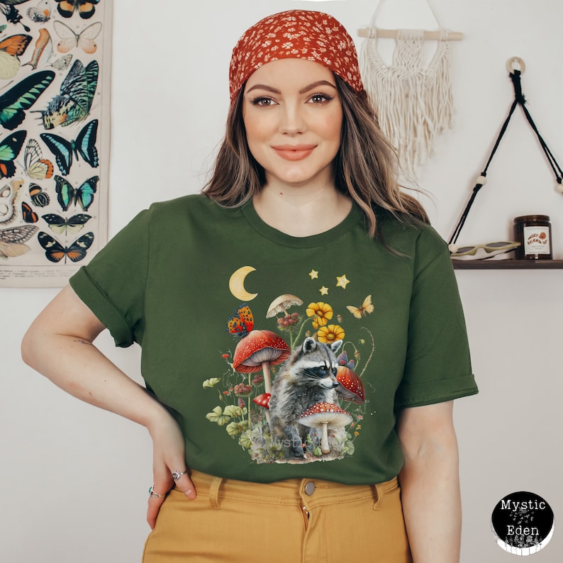 May include: A green t-shirt with a graphic of a raccoon standing in a forest scene with mushrooms, butterflies, and stars. The raccoon is looking to the right. The shirt has a crew neck and short sleeves.
