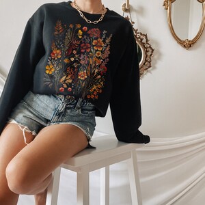 May include: A black sweatshirt with a floral print featuring a variety of dried flowers in shades of orange, yellow, and red. The sweatshirt is being worn by a person sitting on a white stool.