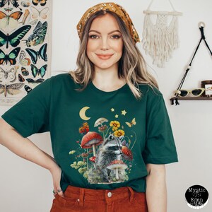 May include: A green t-shirt with a raccoon sitting on a mushroom in a forest scene. The raccoon is surrounded by mushrooms, flowers, and butterflies. The shirt has a crescent moon and stars in the top left corner.