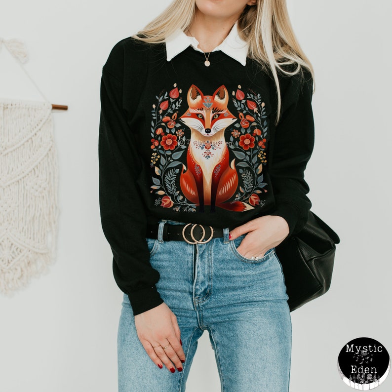 Op de afbeelding: Zwart sweatshirt met een kleurrijke illustratie van een vos omringd door bloemen. De vos draagt een ketting met een geometrisch patroon. Het sweatshirt heeft een witte kraag.