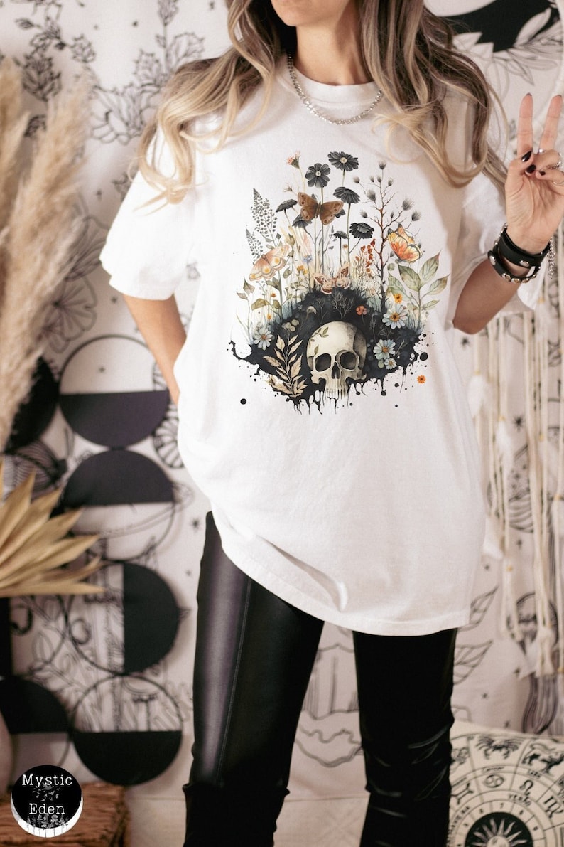 K&ouml;nnte beinhalten: Wei&szlig;es T-Shirt mit einem Aquarelldesign eines Sch&auml;dels, umgeben von Blumen und Schmetterlingen. Die Grafik enth&auml;lt schwarze, orange und gr&uuml;ne Elemente. Das Shirt hat kurze &Auml;rmel und eine lockere Passform.