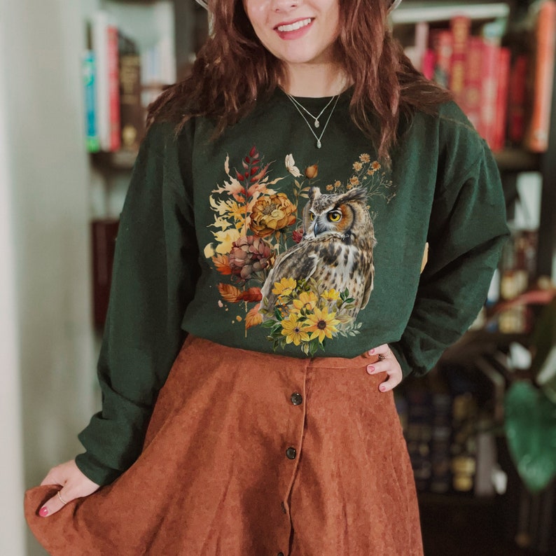 May include: A dark green sweatshirt with a graphic print of an owl surrounded by fall foliage. The owl is brown and gray with yellow eyes. The foliage is a mix of yellow, orange, and brown.