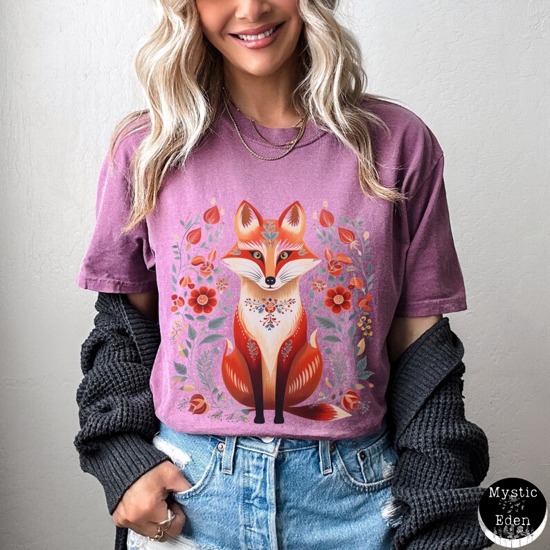 May include: A mauve t-shirt with a detailed illustration of a fox and floral design. The fox is orange and white, with intricate patterns. The surrounding floral elements include red and green accents, creating a vibrant and artistic design.