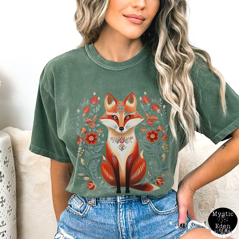 May include: A green t-shirt with a detailed illustration of a fox with red and white accents, surrounded by a floral wreath. The fox has a decorative pattern on its chest. The text "Mystic Eden" is in the lower right corner.