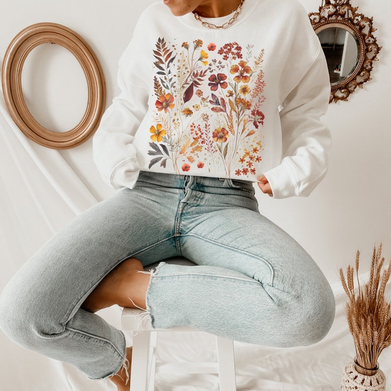 May include: White sweatshirt with a floral print featuring orange, yellow, and red flowers. The flowers are arranged in a vertical pattern on the front of the sweatshirt.