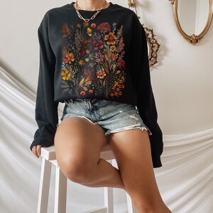 May include: A black sweatshirt with a floral print featuring a variety of flowers in shades of orange, yellow, and red. The flowers are arranged in a bouquet-like design and are set against a black background.
