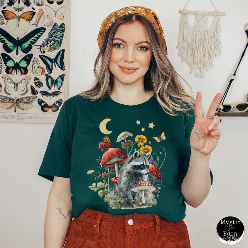 May include: A dark green t-shirt with a graphic of a raccoon in a forest scene with mushrooms, flowers, and a crescent moon. The raccoon is wearing a red bandana.