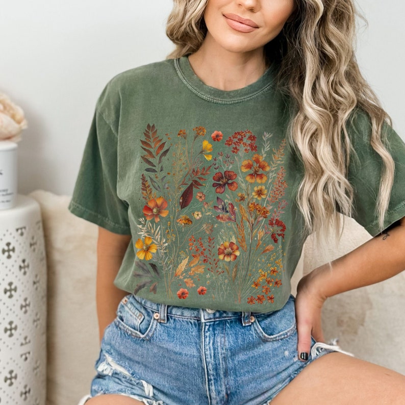 Op de afbeelding: Een groen T-shirt met een bloemenprint met verschillende bloemen in tinten oranje, geel en rood. De bloemen zijn in een boeketvormig patroon op een groene achtergrond gerangschikt.