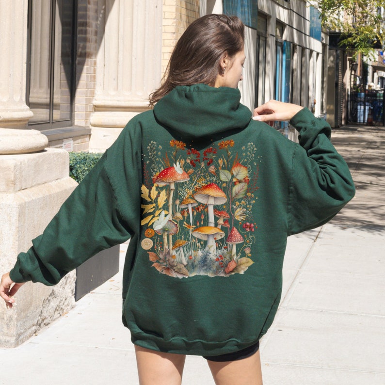 Op de afbeelding: Een donkergroen hoodie met een kleurrijke paddenstoel illustratie op de rug. De illustratie toont rode en witte paddenstoelen met bruine stelen en groene bladeren.