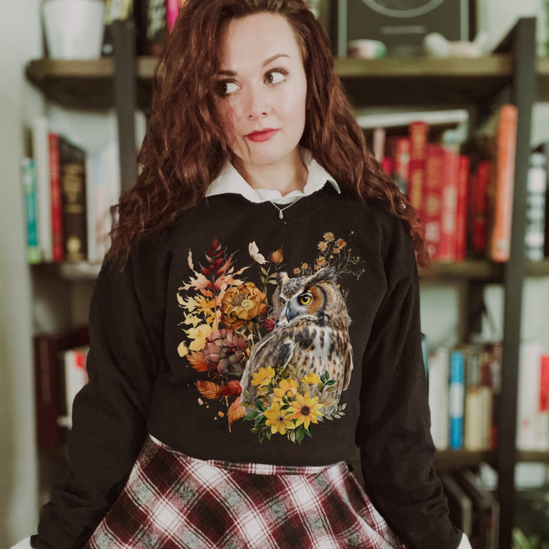 May include: A black cropped sweatshirt with a graphic print of an owl surrounded by fall foliage and flowers. The owl has yellow eyes and brown and white feathers.