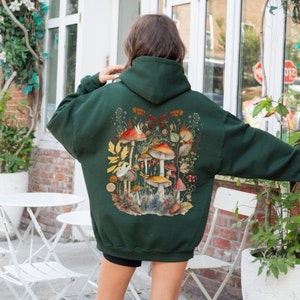 Op de afbeelding: Een donkergroene hoodie met een kleurrijke paddenstoelenillustratie op de rug. De illustratie toont verschillende paddenstoelen met rode hoeden en witte stippen, omringd door bladeren en bloemen.