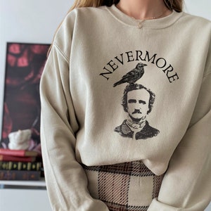 May include: A beige sweatshirt with a black graphic print of a raven perched on the head of Edgar Allan Poe. The text "NEVERMORE" is printed in a black arc above the raven.