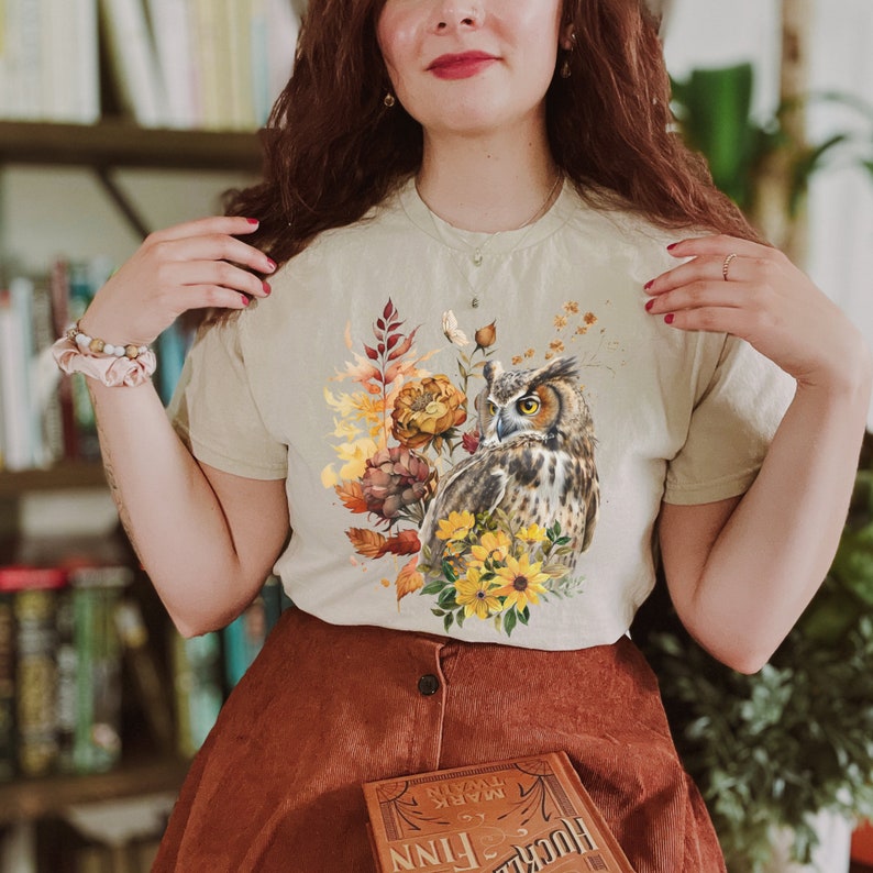 May include: A beige t-shirt with a graphic print of an owl perched on a branch with fall foliage and flowers. The owl has brown and white feathers and yellow eyes. The flowers are yellow, orange, and red.