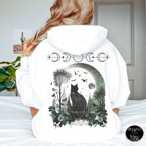 May include: White hooded sweatshirt with a black cat silhouette in a moonlit forest scene. The cat is surrounded by trees, flowers, and birds. The moon phases are depicted above the cat with a star on each side. The text "The Mystic Eden" is visible at the bottom of the design.