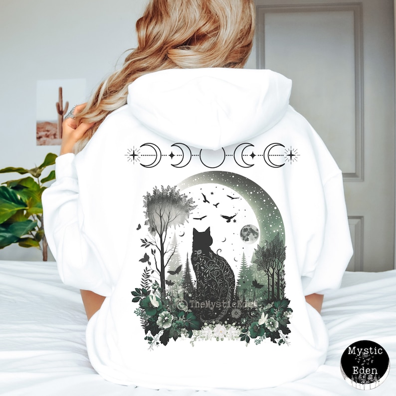 May include: White hooded sweatshirt with a black cat silhouette in a moonlit forest scene. The cat is surrounded by trees, flowers, and birds. The moon phases are depicted above the cat with a star on each side. The text "The Mystic Eden" is visible at the bottom of the design.