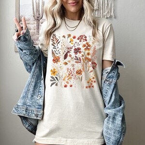 Puede incluir: Una camiseta de color crema con un estampado floral con flores naranjas, rojas y amarillas. Las flores est&aacute;n dispuestas en un patr&oacute;n cuadrado en la parte delantera de la camiseta.