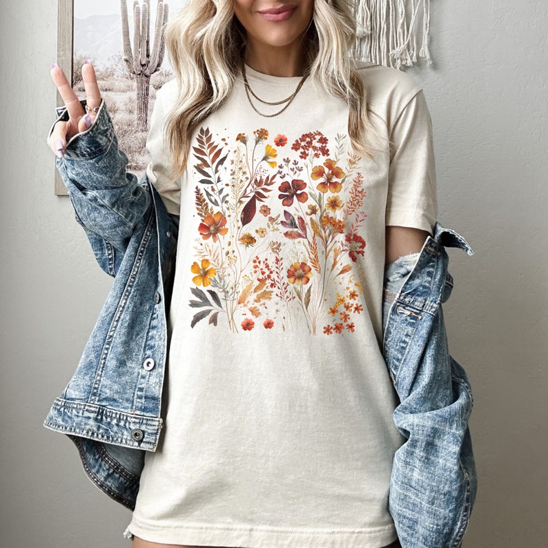 Puede incluir: Una camiseta de color crema con un estampado floral con flores naranjas, rojas y amarillas. Las flores est&aacute;n dispuestas en un patr&oacute;n cuadrado en la parte delantera de la camiseta.