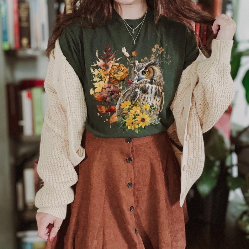 May include: A green t-shirt with a floral print featuring an owl and fall colors. The owl is brown and white with yellow eyes. The flowers are yellow, orange, and red. The shirt is paired with a brown corduroy skirt.