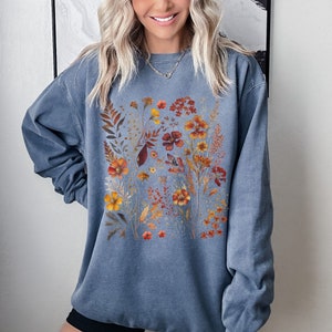 May include: A blue-grey sweatshirt featuring a floral design with orange, yellow, and red flowers and foliage. The botanical print is centered on the front of the sweatshirt, creating a visually appealing contrast against the solid-colored fabric.