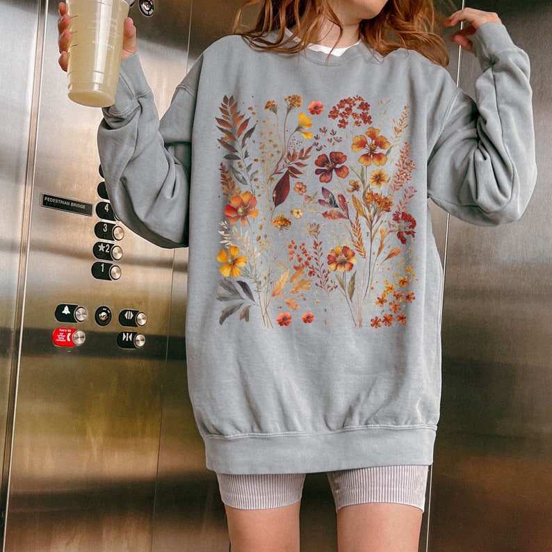 May include: A light blue sweatshirt featuring a floral design with orange, yellow, and red flowers. The sweatshirt has a crew neck and long sleeves. The floral design is centered on the front of the sweatshirt.
