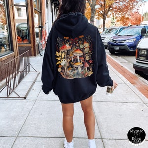 Op de afbeelding: Zwart hoodie met een kleurrijke paddenstoelprint op de rug. De print toont een groep paddenstoelen met rode, oranje en gele hoeden, omringd door bladeren en bloemen.