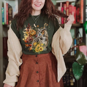 May include: A green t-shirt with a brown and white owl surrounded by fall foliage and flowers. The owl is looking to the left. The shirt is being worn by a person with brown hair and a brown skirt.