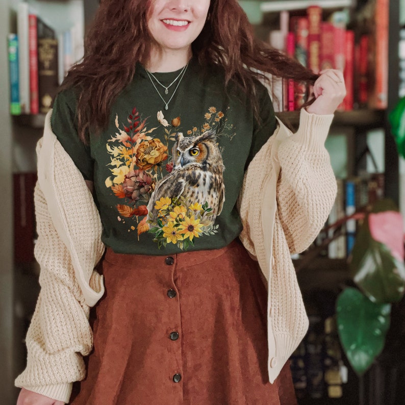 May include: A green t-shirt with a brown and white owl surrounded by fall foliage and flowers. The owl is looking to the left. The shirt is being worn by a person with brown hair and a brown skirt.