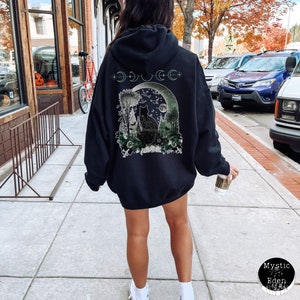 May include: Black hooded sweatshirt with a graphic print of a black cat sitting in a forest under a crescent moon. The graphic also includes a crescent moon, stars, and the text "333 CE".