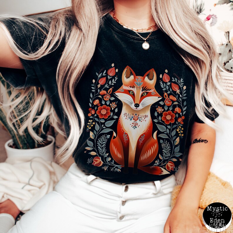 May include: Black t-shirt with a fox illustration. The fox is in orange, brown, and white, surrounded by red, blue, and green floral and leaf designs. The shirt has a crew neck and short sleeves. The design is centered on the chest.