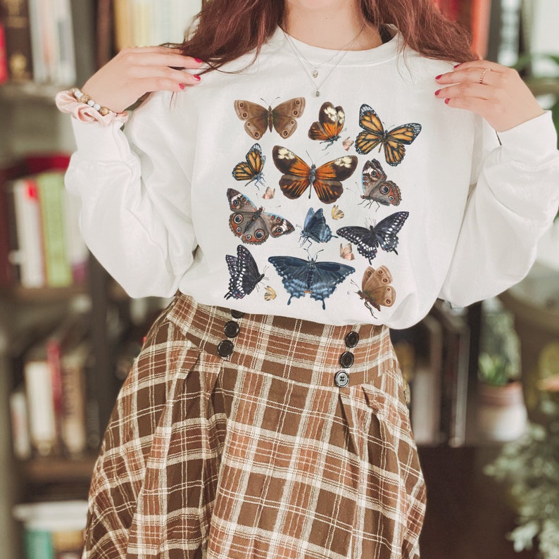 May include: White sweatshirt with a colorful butterfly print. The butterflies are in various shades of brown, orange, and blue. The sweatshirt is paired with a brown and white plaid skirt.
