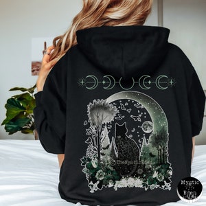 May include: Black hoodie with a graphic print of a cat sitting in a forest under a crescent moon. The moon is surrounded by stars and the graphic includes the text "The Mystic Eden".
