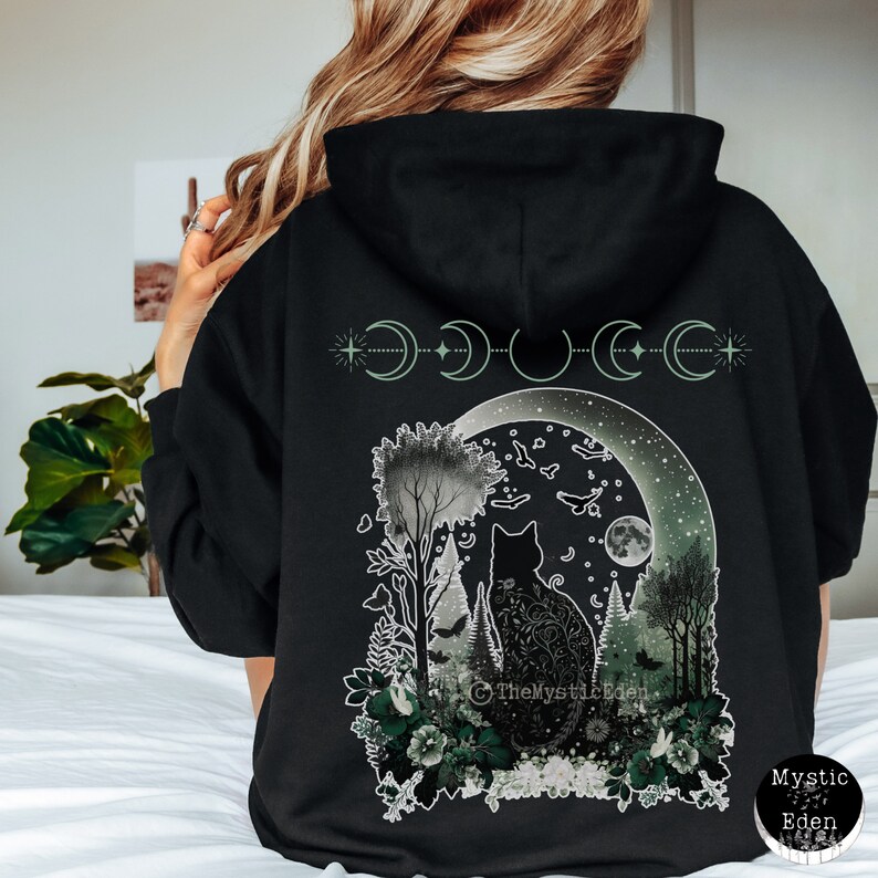 May include: Black hoodie with a graphic print of a cat sitting in a forest under a crescent moon. The moon is surrounded by stars and the graphic includes the text "The Mystic Eden".