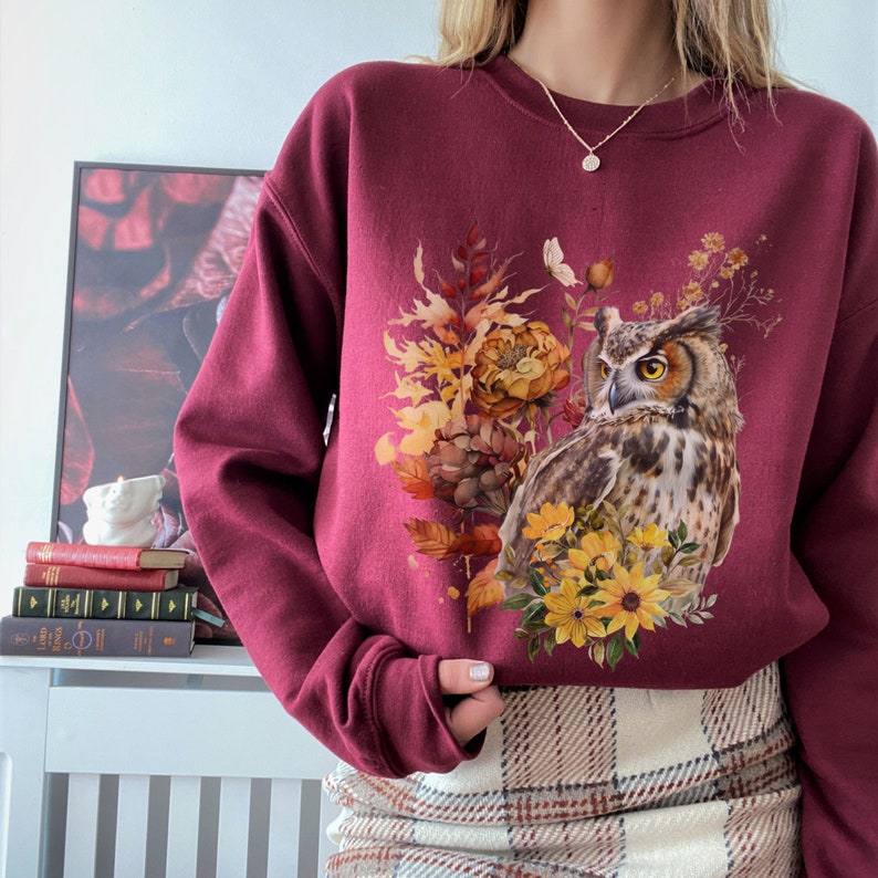 May include: A maroon sweatshirt with a graphic print of an owl surrounded by fall foliage and flowers. The owl is brown and white with yellow eyes. The flowers are yellow and orange.