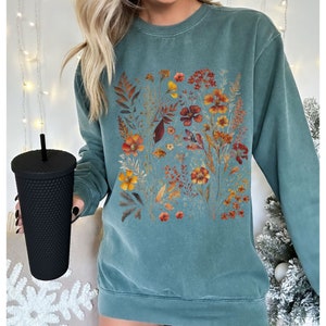 May include: Teal-colored sweatshirt featuring a floral design with orange, yellow, and red flowers and foliage. The design is centered on the front of the sweatshirt. The sweatshirt is a crew neck style and has long sleeves. A black textured tumbler with a straw is held in the image.