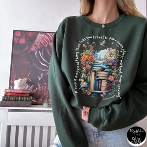 K&ouml;nnte beinhalten: Ein dunkelgr&uuml;ner Pullover mit einem Zitat &uuml;ber B&uuml;cher, das auf der Vorderseite aufgedruckt ist. Das Zitat lautet: "Ein Buch ist eine magische Sache, die es dir erm&ouml;glicht, an ferne Orte zu reisen, ohne jemals zu gehen."