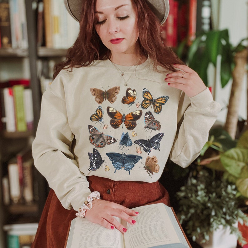 May include: A beige sweatshirt with a colorful butterfly print. The butterflies are in various shades of blue, green, orange, and brown. The sweatshirt has long sleeves and a crew neck.