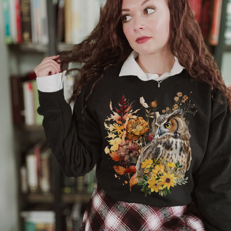 May include: Black sweatshirt with a graphic print of an owl surrounded by fall foliage and flowers. The owl has brown and white feathers and yellow eyes.