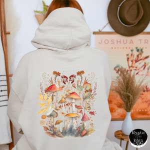 Op de afbeelding: Een beige hoodie met een kleurrijk ontwerp van paddenstoelen en bloemen op de rug. Het ontwerp bevat rode, oranje, gele en bruine paddenstoelen met groene bladeren en bloemen.
