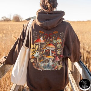 Op de afbeelding: Bruin hoodie met een kleurrijk ontwerp van paddenstoelen en bladeren op de rug. Het ontwerp bevat een verscheidenheid aan paddenstoelen, bladeren en bloemen in tinten rood, oranje, geel, groen en bruin.