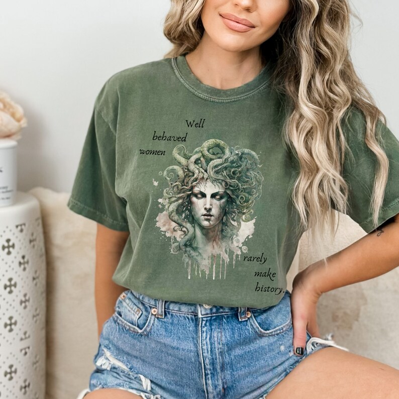 May include: Olive green t-shirt featuring a watercolor illustration of Medusa with text that reads "Well behaved women rarely make history." The graphic is in shades of green, gray, and white. The shirt has a relaxed fit.