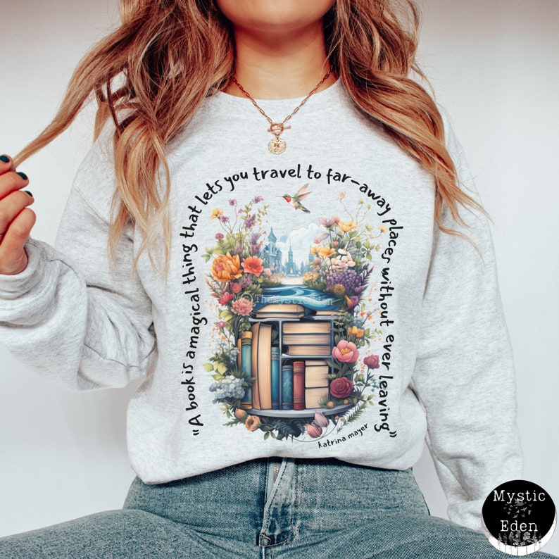 K&ouml;nnte beinhalten: Ein graues Sweatshirt mit einem grafischen Druck eines B&uuml;cherregals gef&uuml;llt mit B&uuml;chern, Blumen und einem Kolibri. Der Text "A book is a magical thing that lets you travel to far-away places without ever leaving" ist auf dem Sweatshirt gedruckt.