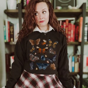 May include: A black cropped sweatshirt with a colorful butterfly print. The butterflies are arranged in a random pattern and are of various sizes and colors.