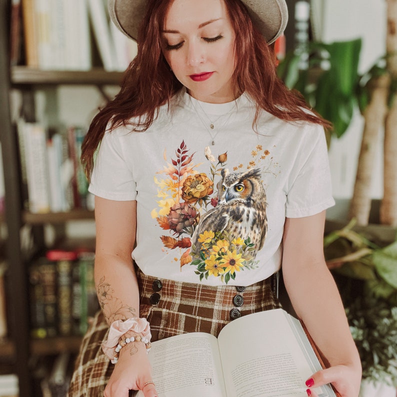 May include: White t-shirt with a watercolor design of an owl, flowers, and leaves in shades of brown, yellow, and orange.