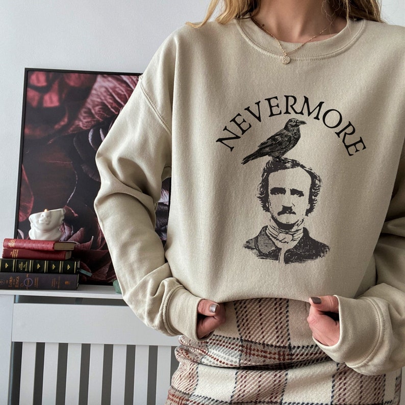 May include: A beige sweatshirt with a black graphic print of Edgar Allan Poe with a raven perched on his head. The text "NEVERMORE" is printed above the graphic.