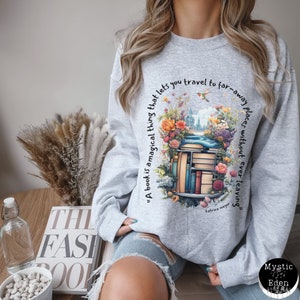 K&ouml;nnte beinhalten: Ein graues Sweatshirt mit einem grafischen Druck eines Stapels B&uuml;cher, umgeben von Blumen und einem Zitat, das lautet: "Ein Buch ist eine magische Sache, die es dir erm&ouml;glicht, an ferne Orte zu reisen, ohne jemals wegzugehen" von Katrina Mayer.